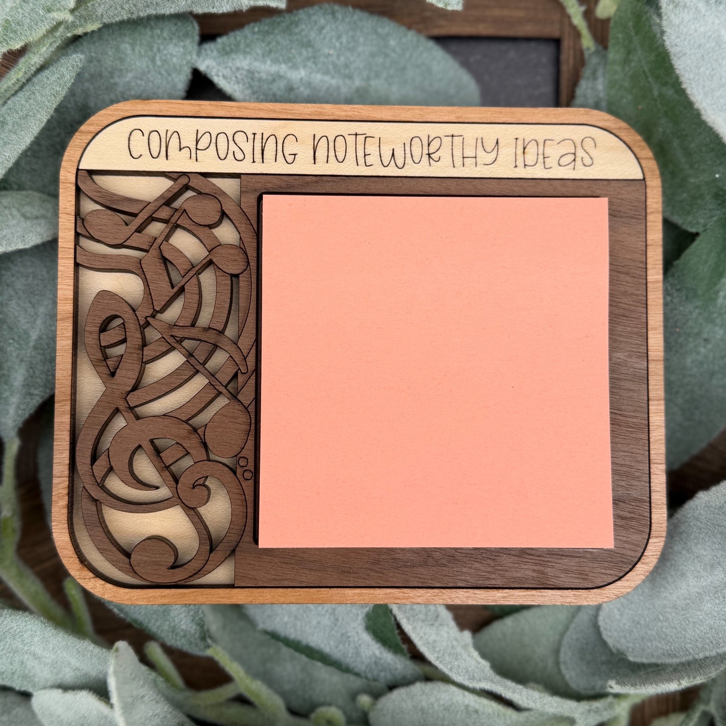 Wooden notepad with decorative design and text 'Composing Noteworthy Ideas' on a leafy background
