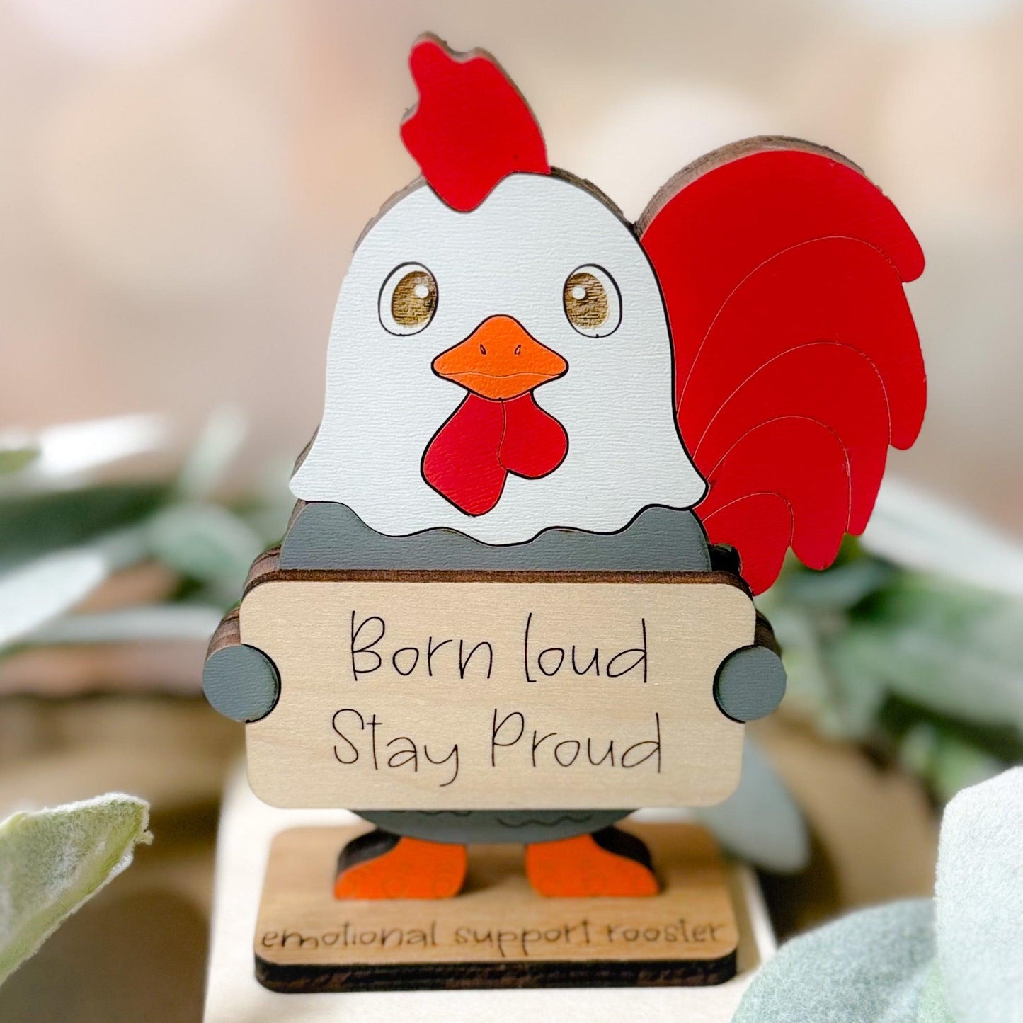Decorative rooster figurine with 'Born loud Stay Proud' message on a blurred natural background