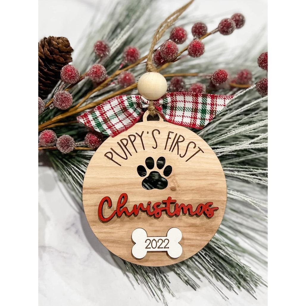 Wooden Puppy’s First Christmas ornament with paw cut-out, red “Christmas” lettering, engraved bone with 2022, plaid bow, and rustic twine hanger on holiday greenery.