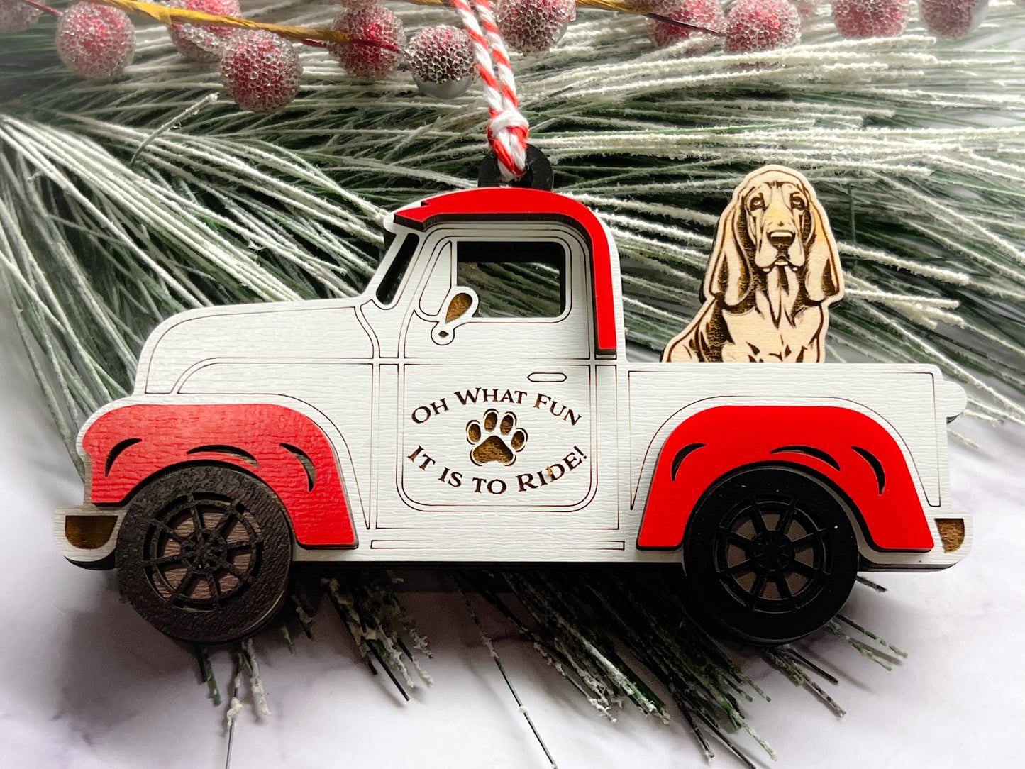 A vintage style red and white truck ornament with a dog riding in the back seat, placed on a branch with Christmas decorations.