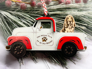 A vintage style red and white truck ornament with a dog riding in the back seat, placed on a branch with Christmas decorations.