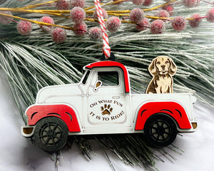 A vintage style red and white truck ornament with a dog riding in the back seat, placed on a branch with Christmas decorations.