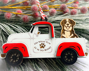 A vintage style red and white truck ornament with a dog riding in the back seat, placed on a branch with Christmas decorations.