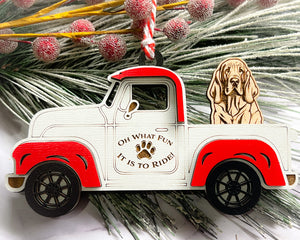 A vintage style red and white truck ornament with a dog riding in the back seat, placed on a branch with Christmas decorations.