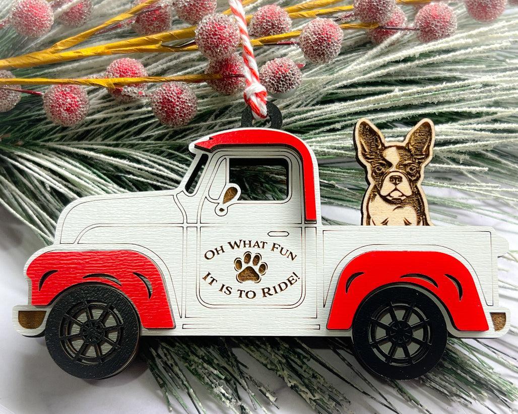 A vintage style red and white truck ornament with a dog riding in the back seat, placed on a branch with Christmas decorations.