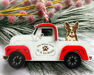 A vintage style red and white truck ornament with a dog riding in the back seat, placed on a branch with Christmas decorations.