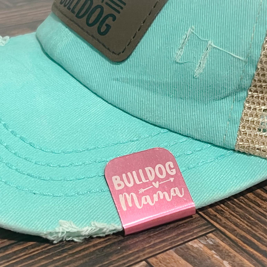 Close-up of a Bulldog Mama metal hat clip attached to the brim of a teal and tan ball cap on a wooden surface.