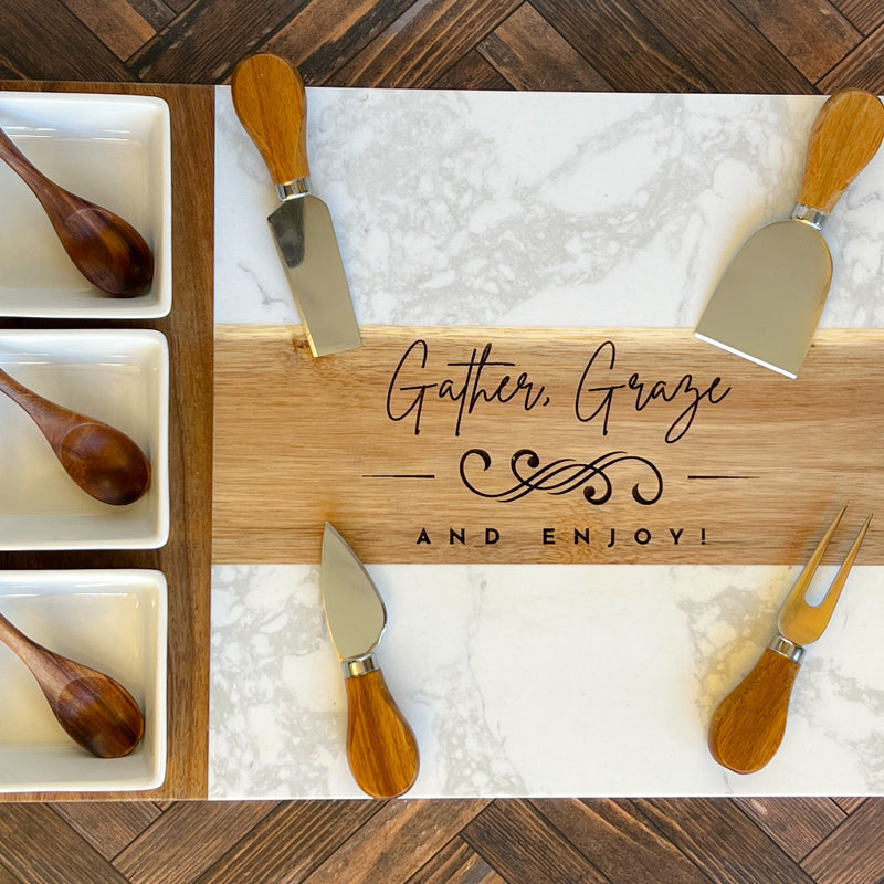 Engraved marble and wood charcuterie board with phrase “Gather, Graze, and Enjoy,” displayed with cheese knives, fork, and wooden spoons in ceramic bowls.