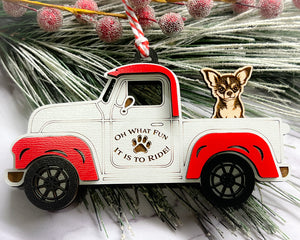 A vintage style red and white truck ornament with a dog riding in the back seat, placed on a branch with Christmas decorations.