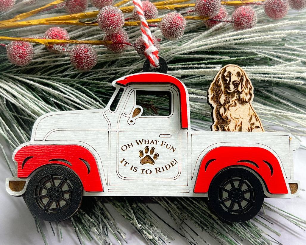 A vintage style red and white truck ornament with a dog riding in the back seat, placed on a branch with Christmas decorations.
