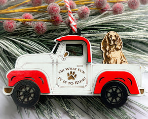A vintage style red and white truck ornament with a dog riding in the back seat, placed on a branch with Christmas decorations.