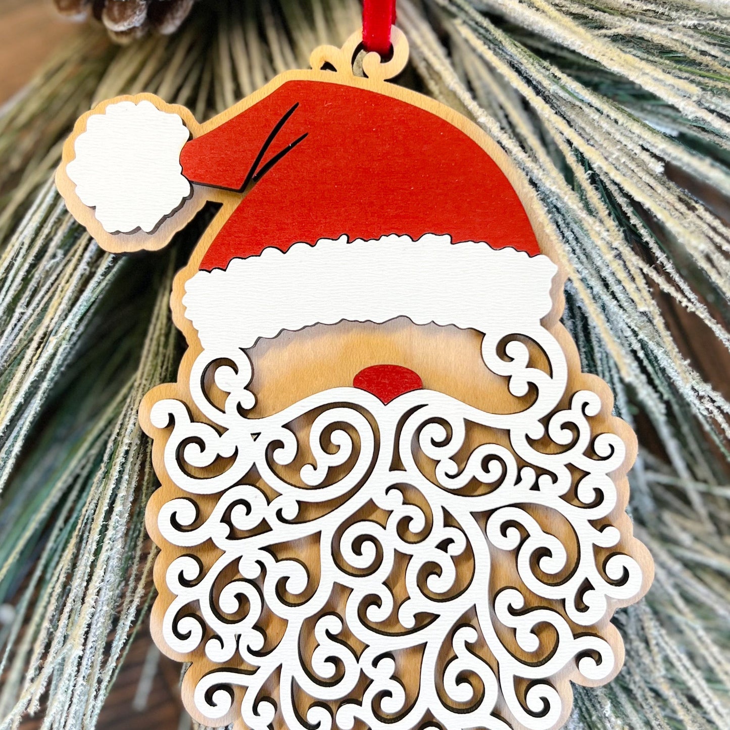 A laser-cut wood Santa ornament with a red velour ribbon and a curly beard design, placed on a branch with pine needles.