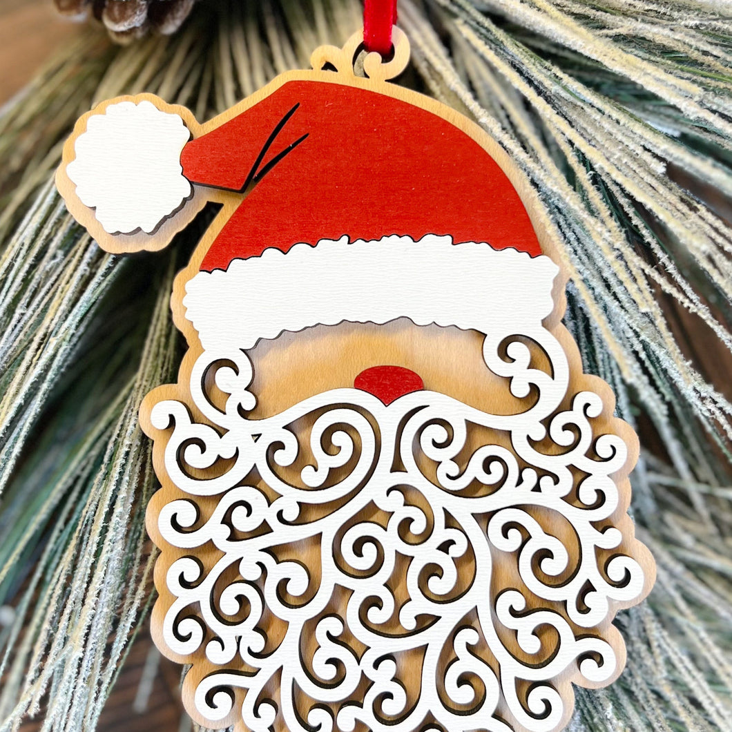 A laser-cut wood Santa ornament with a red velour ribbon and a curly beard design, placed on a branch with pine needles.