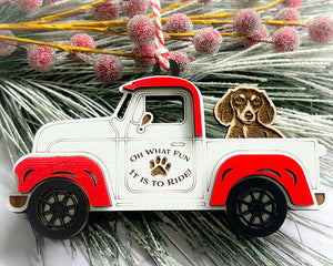 A vintage style red and white truck ornament with a dog riding in the back seat, placed on a branch with Christmas decorations.