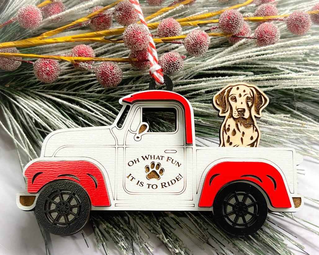 A vintage style red and white truck ornament with a dog riding in the back seat, placed on a branch with Christmas decorations.