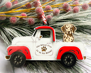 A vintage style red and white truck ornament with a dog riding in the back seat, placed on a branch with Christmas decorations.