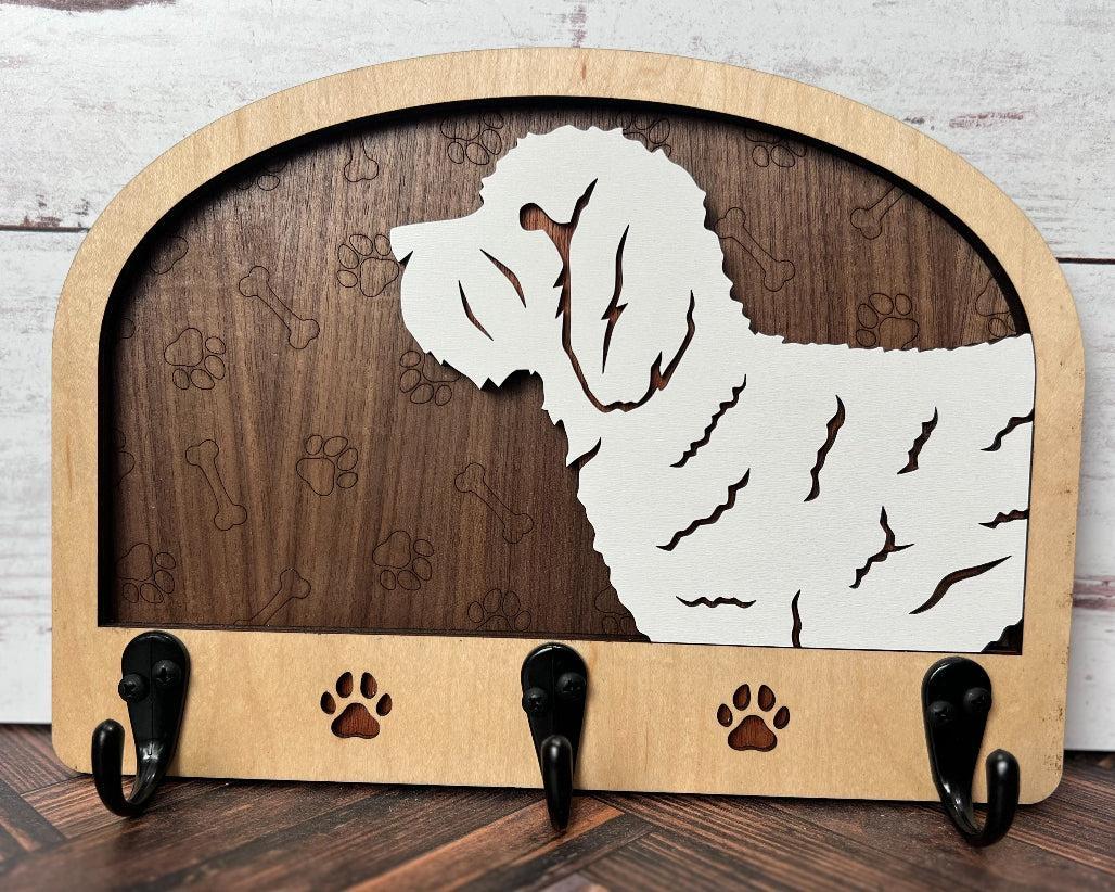 A wooden dog leash holder with a cut-out silhouette of a dog and the phrase 'Unleash Adventure'. It features three hooks at the bottom and paw prints around the dog's silhouette.