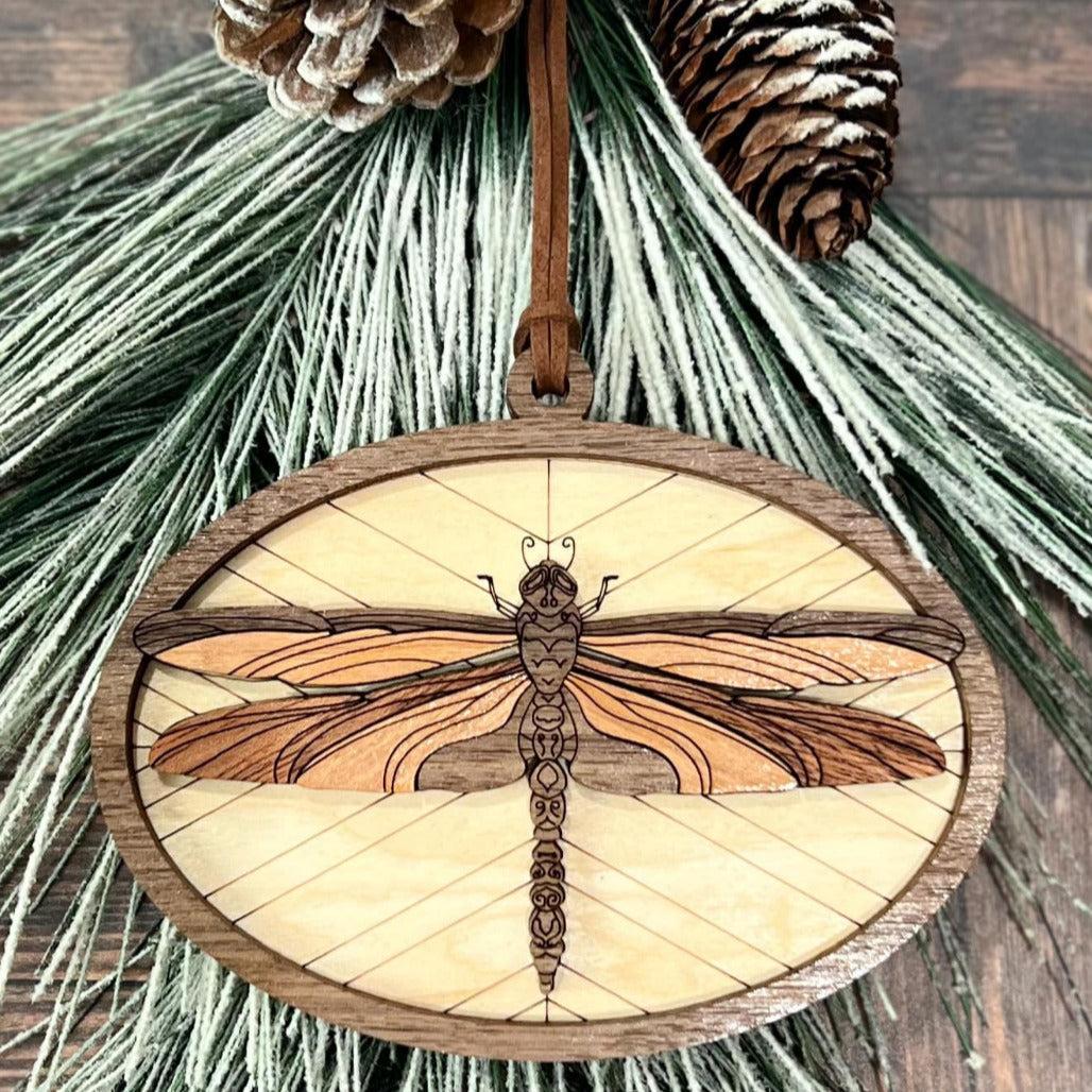 A circular wooden ornament featuring a laser-cut dragonfly design, with a pine cone backdrop.