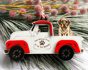 A vintage style red and white truck ornament with a dog riding in the back seat, placed on a branch with Christmas decorations.