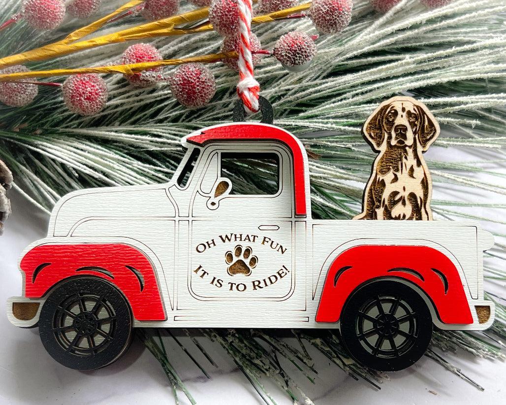 A vintage style red and white truck ornament with a dog riding in the back seat, placed on a branch with Christmas decorations.