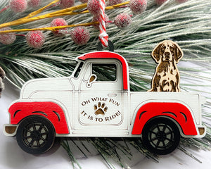 A vintage style red and white truck ornament with a dog riding in the back seat, placed on a branch with Christmas decorations.