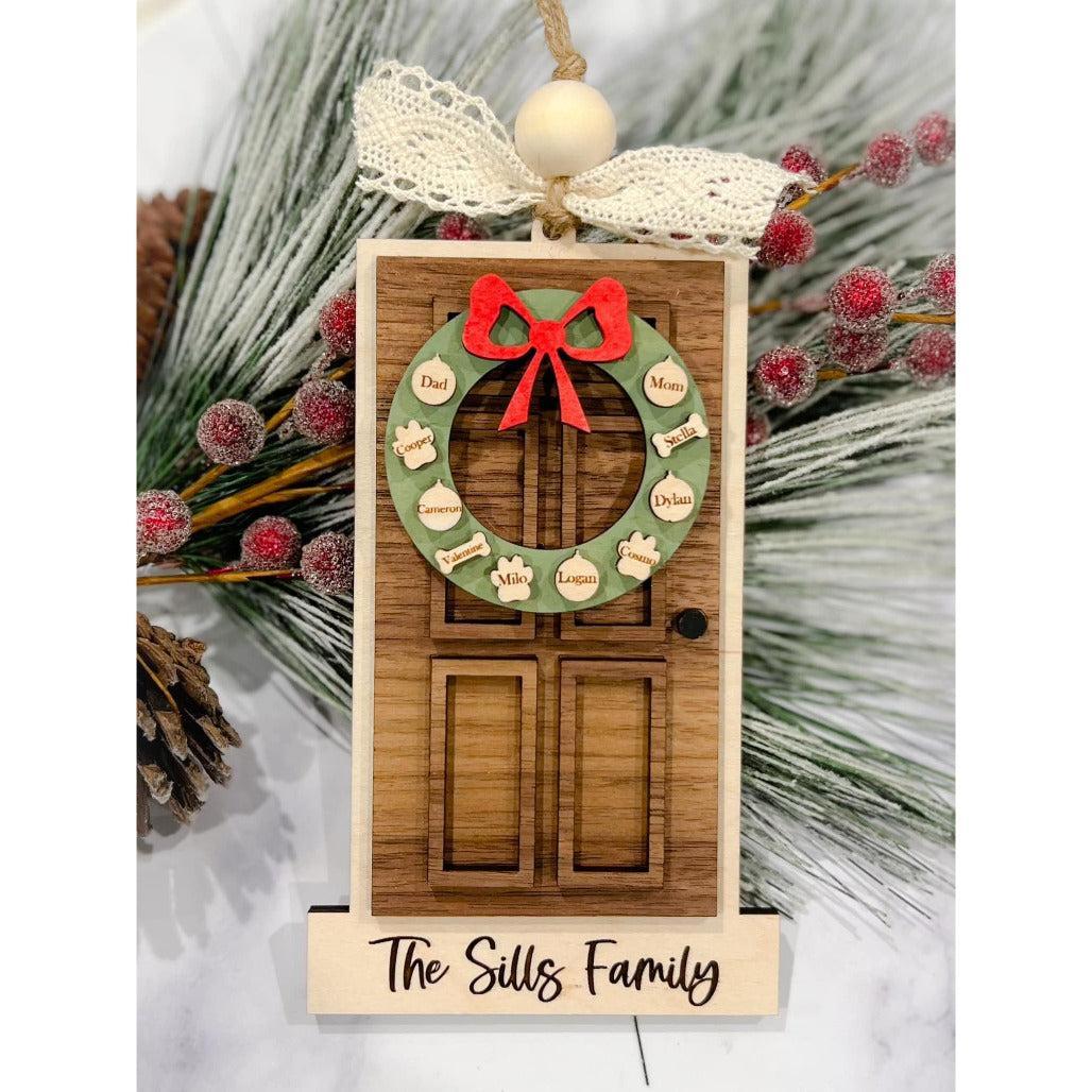 Wooden family door ornament with engraved names on a wreath and “The Sills Family” at the bottom, decorated with lace bow, pinecones, and frosted greenery.