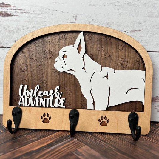 A wooden dog leash holder with a cut-out silhouette of a dog and the phrase 'Unleash Adventure'. It features three hooks at the bottom and paw prints around the dog's silhouette.