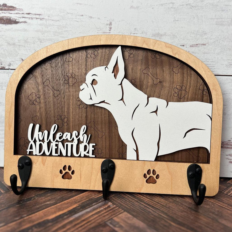 A wooden dog leash holder with a cut-out silhouette of a dog and the phrase 'Unleash Adventure'. It features three hooks at the bottom and paw prints around the dog's silhouette.