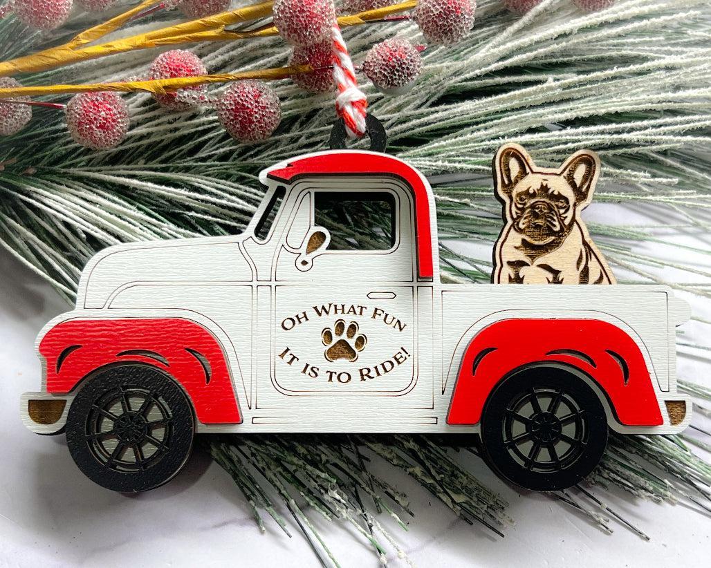 A vintage style red and white truck ornament with a dog riding in the back seat, placed on a branch with Christmas decorations.