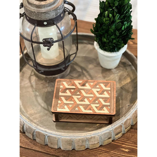 A wooden box with a geometric pattern on the lid, placed on a metal tray, with a candle and a small potted plant in the background.
