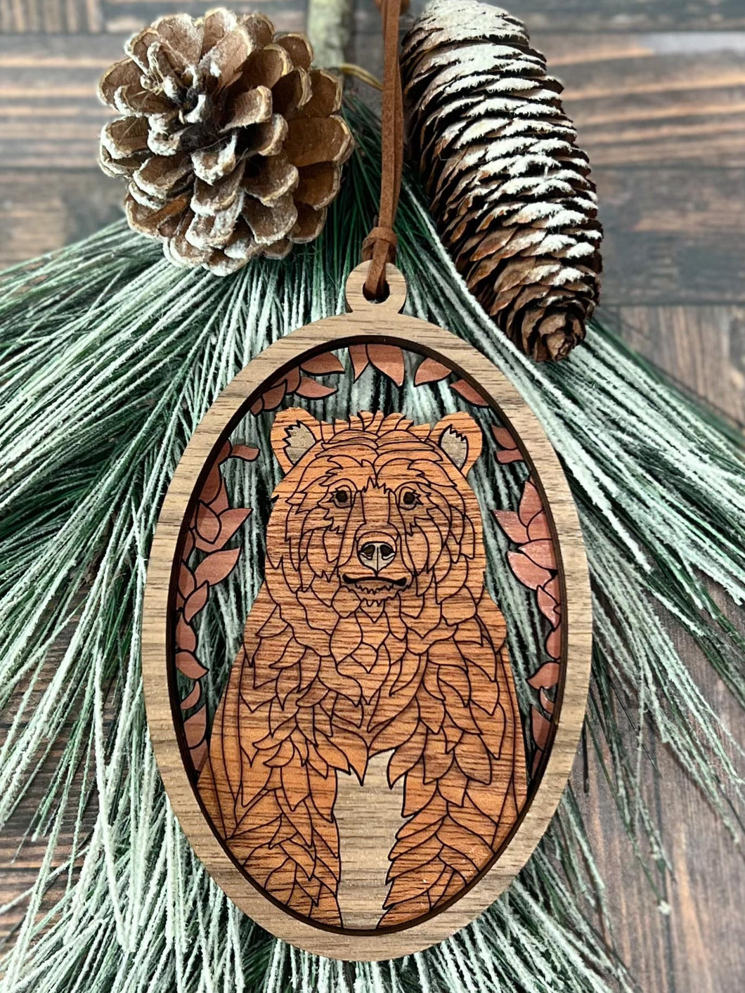 Wooden bear Christmas ornament with layered details and leaf border, hanging on a pine branch with pinecones.