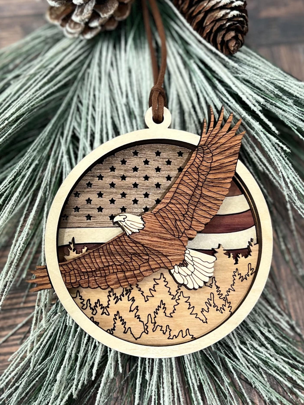 Wooden eagle ornament with layered details, showing a bald eagle in flight over trees with a U.S. flag and stars in the background.