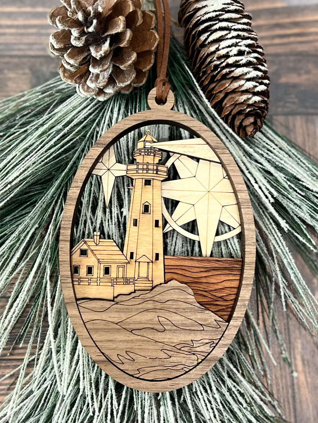 A laser-cut Heirloom Lighthouse Ornament with a wooden frame, featuring an intricate lighthouse design, displayed against a backdrop of green pine needles and a few pine cones.