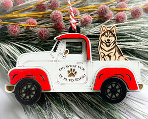 A vintage style red and white truck ornament with a dog riding in the back seat, placed on a branch with Christmas decorations.