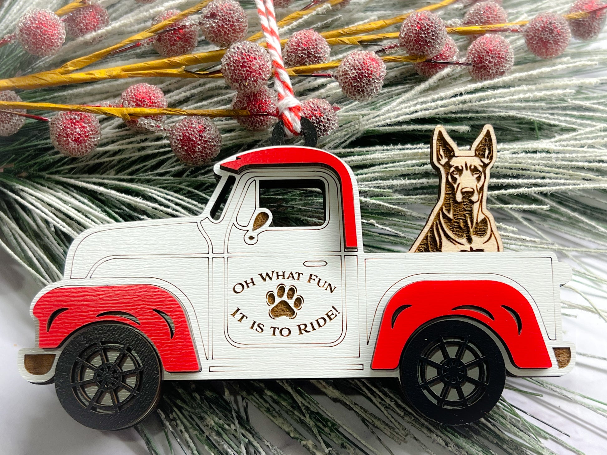 A vintage style red and white truck ornament with a dog riding in the back seat, placed on a branch with Christmas decorations.