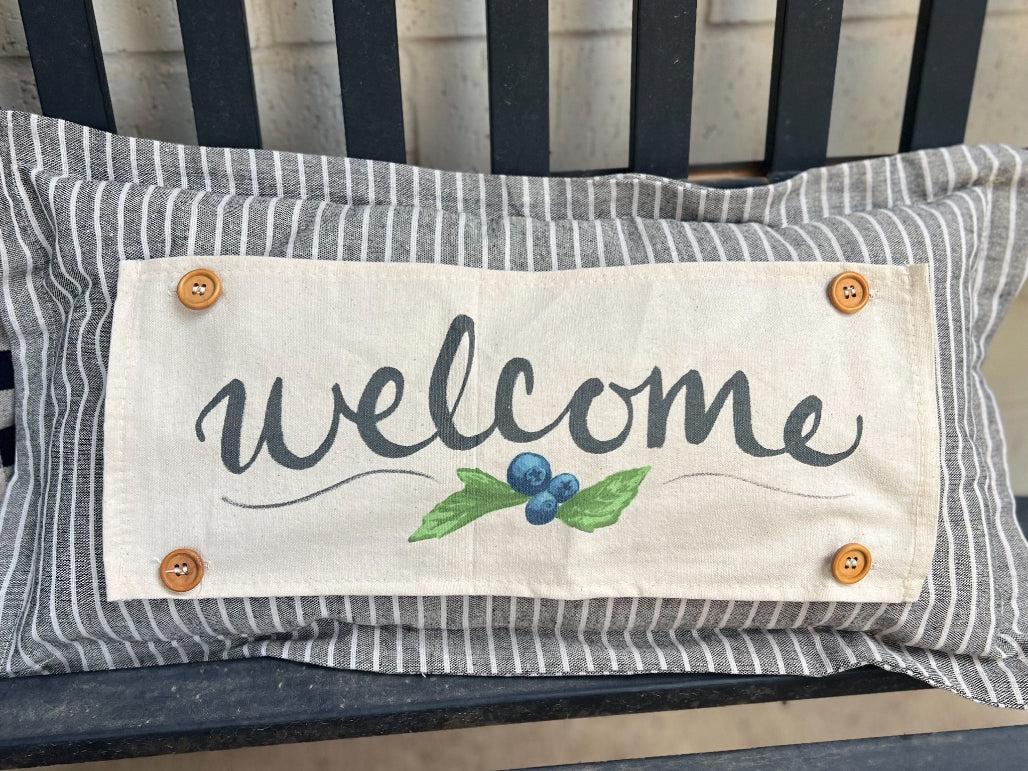 Decorative pillow with 'welcome' text and blueberries on a striped bench.