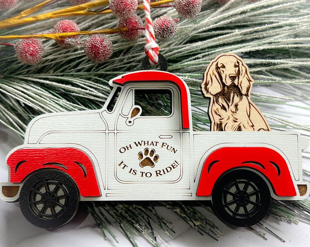 A vintage style red and white truck ornament with a dog riding in the back seat, placed on a branch with Christmas decorations.