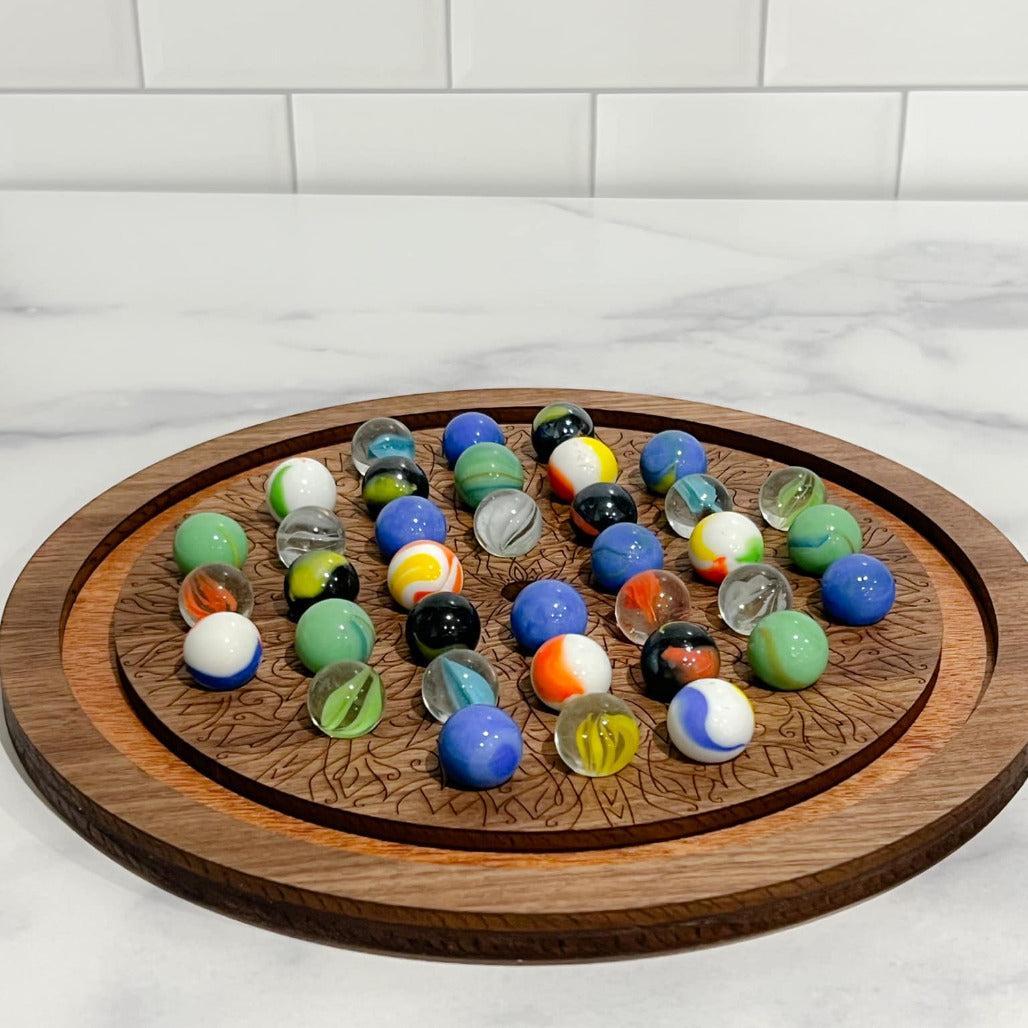 A wooden solitaire marble game with a circular board and various colored marbles placed on it, set against a marble-tiled surface.