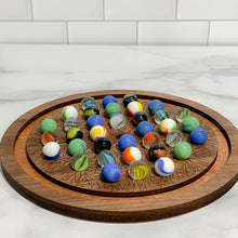 Load image into Gallery viewer, A wooden solitaire marble game with a circular board and various colored marbles placed on it, set against a marble-tiled surface.