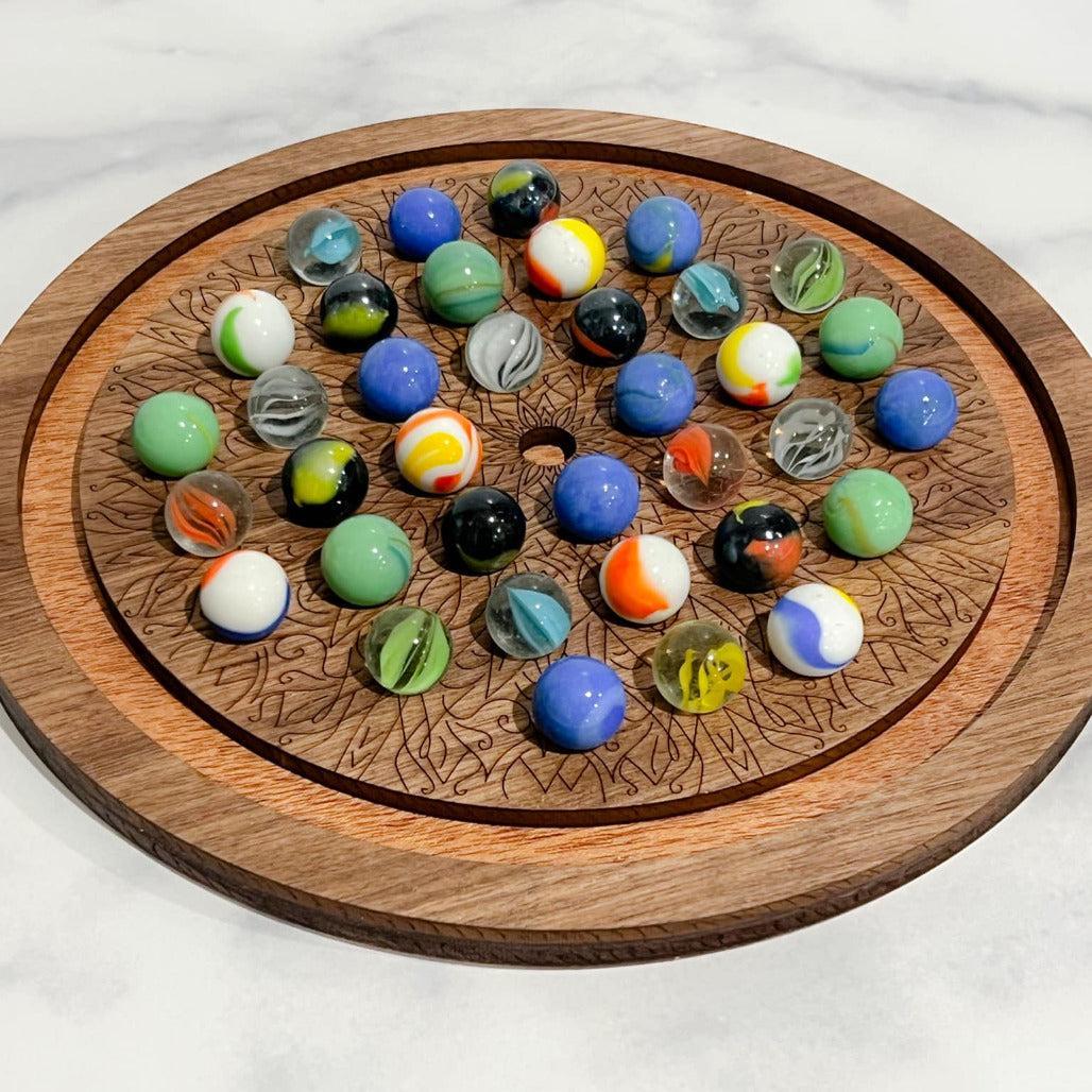 A wooden solitaire marble game with a circular board and various colored marbles placed on it, set against a marble-tiled surface.