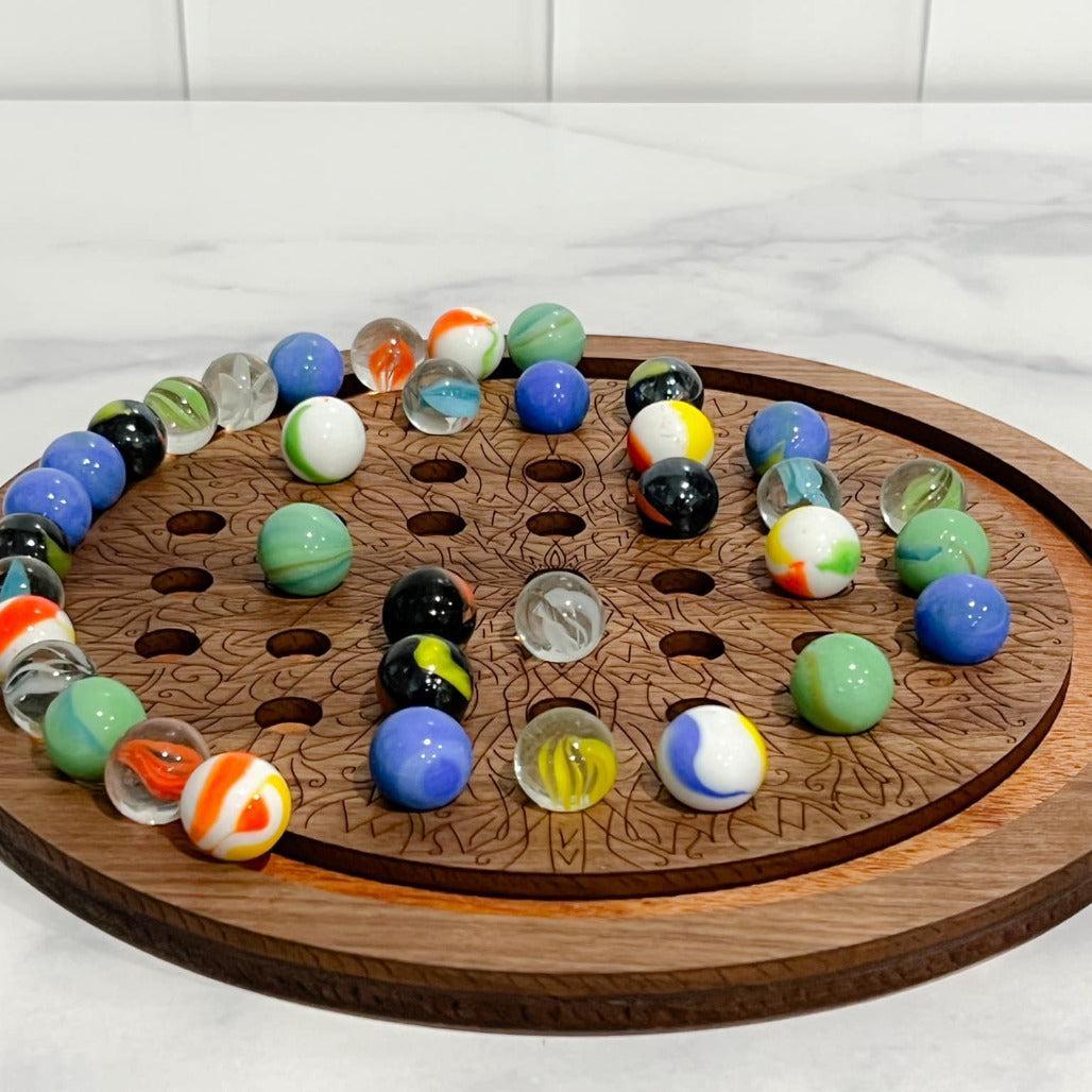 A wooden solitaire marble game with a circular board and various colored marbles placed on it, set against a marble-tiled surface.