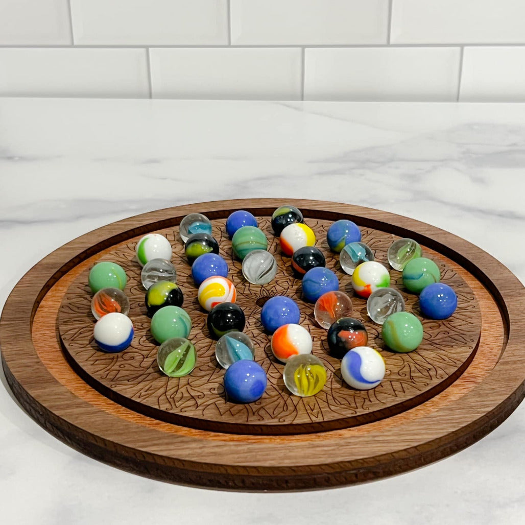 A wooden solitaire marble game with a circular board and various colored marbles placed on it, set against a marble-tiled surface.