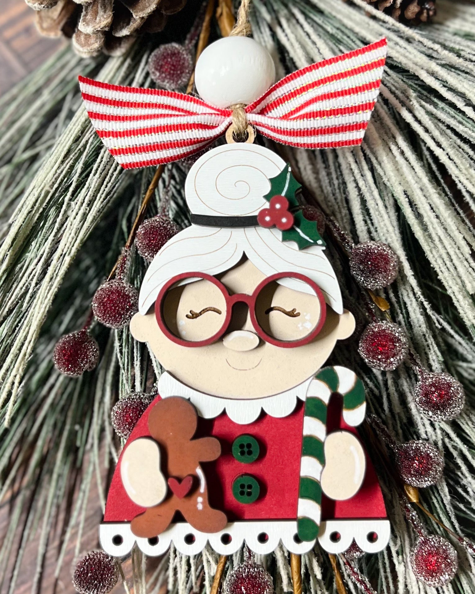 A wooden laser-cut ornament depicting a stylized Mrs Claus with a red dress, green buttons, and a scalloped hem. She is holding a gingerbread cookie and has a red and white striped bow on top for hanging.
