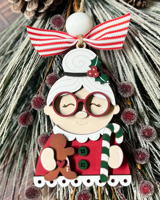 A wooden laser-cut ornament depicting a stylized Mrs Claus with a red dress, green buttons, and a scalloped hem. She is holding a gingerbread cookie and has a red and white striped bow on top for hanging.