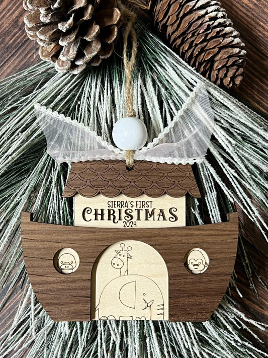 A personalized Noah's Ark ornament made of wood, with a pinecone decoration at the top, a white and pink ribbon, and a 'Christmas' sign in the front.