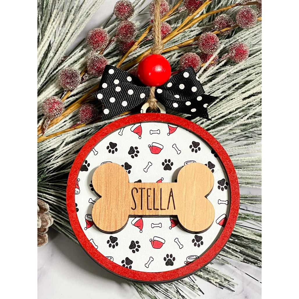 Round pet Christmas ornament with red border, paw print and bone background, wooden bone engraved with “Stella,” polka dot bow, and red bead hanging on frosted greenery.