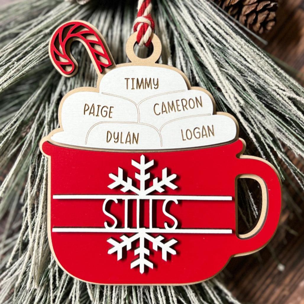 Red hot cocoa mug Christmas ornament with engraved family name “Sills,” marshmallow toppers engraved with names, and candy cane accent hanging on frosted greenery.