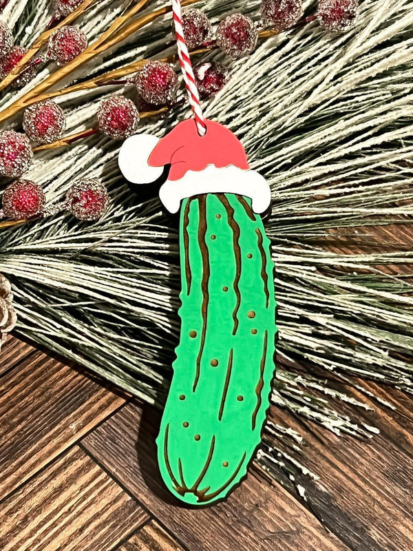 Wooden Christmas pickle ornament painted green with a red Santa hat, hanging on frosted pine branches with red berries.