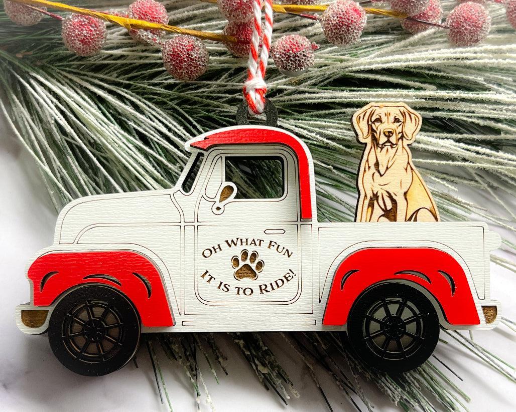 A vintage style red and white truck ornament with a dog riding in the back seat, placed on a branch with Christmas decorations.