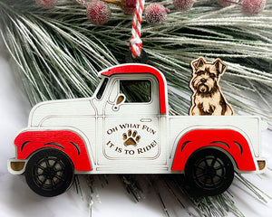 A vintage style red and white truck ornament with a dog riding in the back seat, placed on a branch with Christmas decorations.